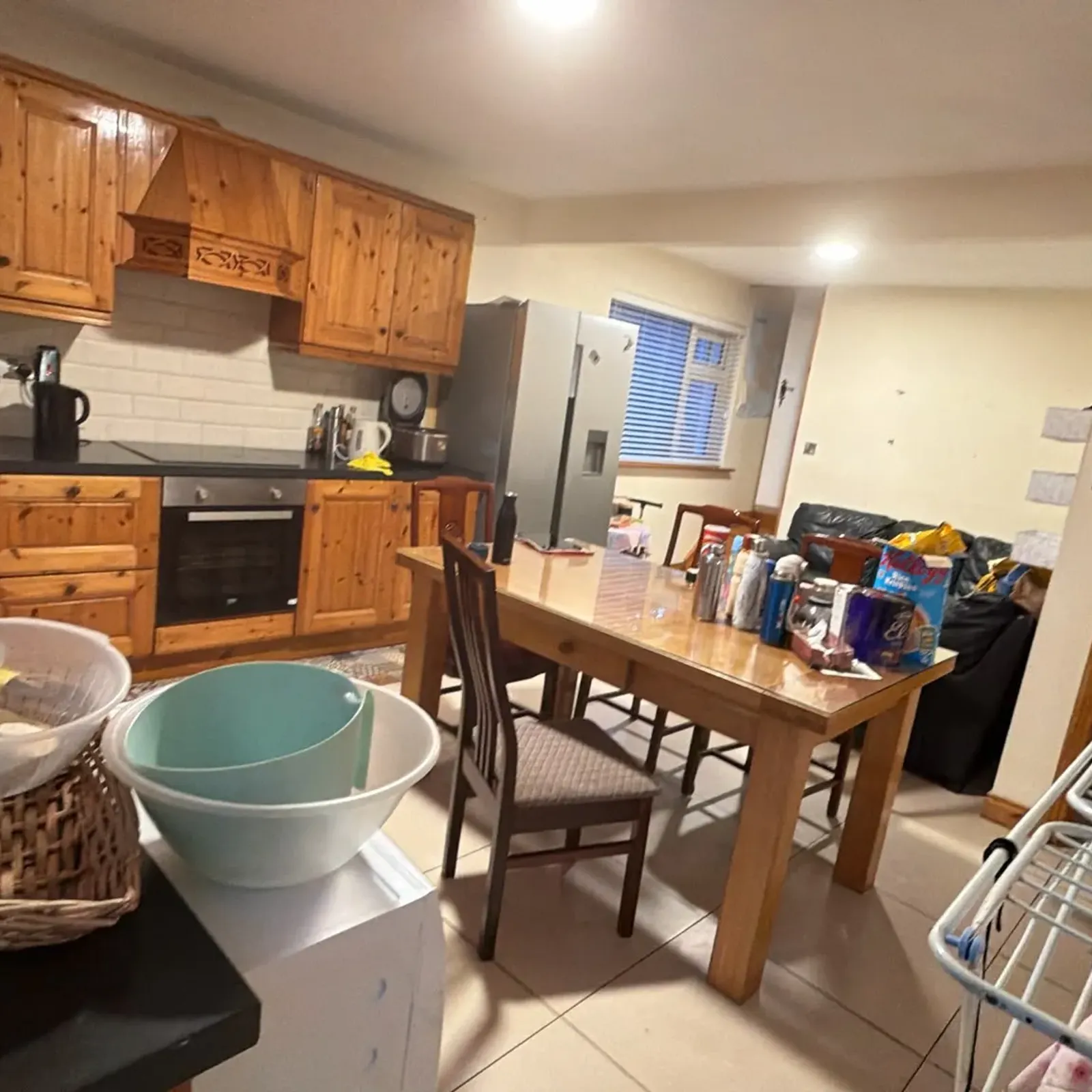 Open-plan kitchen with wooden upper and lower cabinets, built-in oven, stainless refrigerator, sink area and a wooden dining table with chairs and items on the table.
