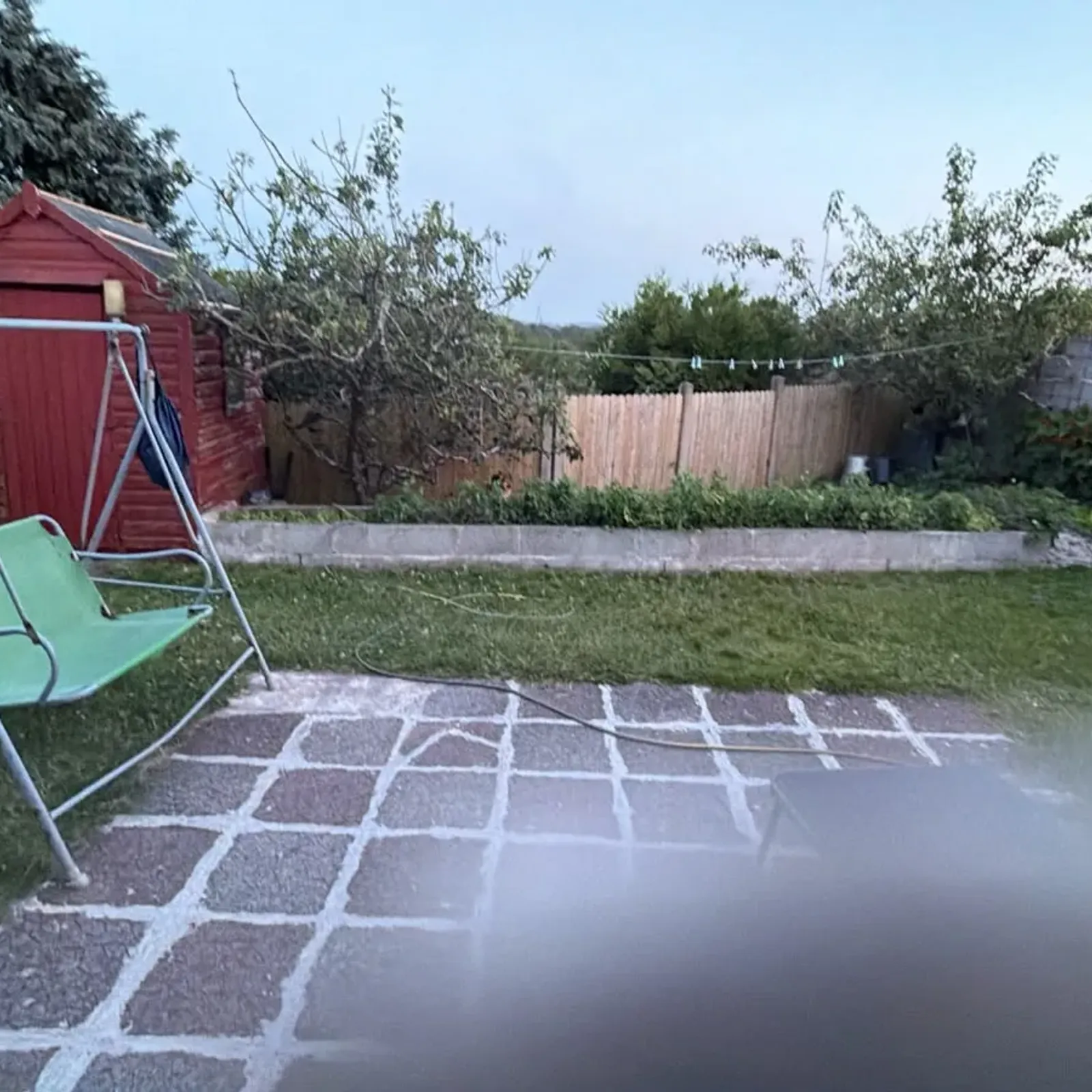 Backyard with a paved patio, lawn, green metal-framed swing chair, red wooden shed, garden beds and a clothesline.