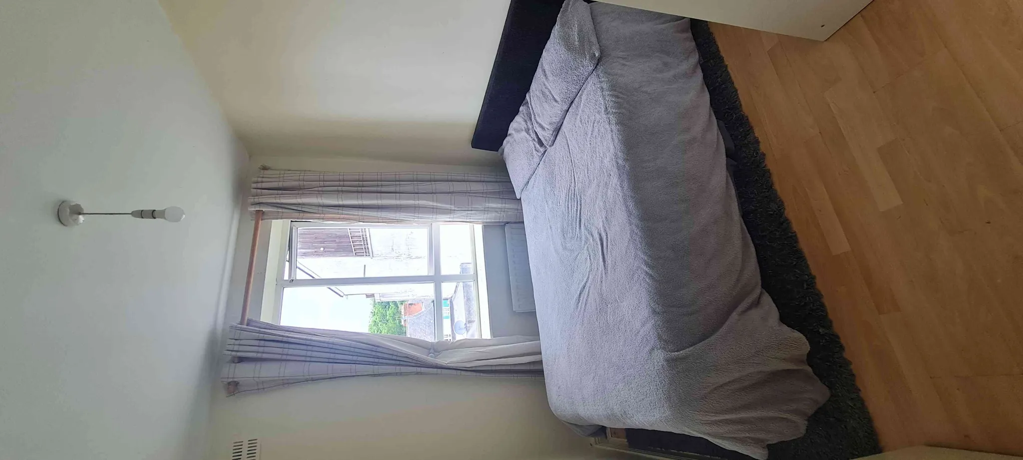 Bedroom with a bed covered by a grey blanket, window with curtains, wooden floor and a ceiling light.