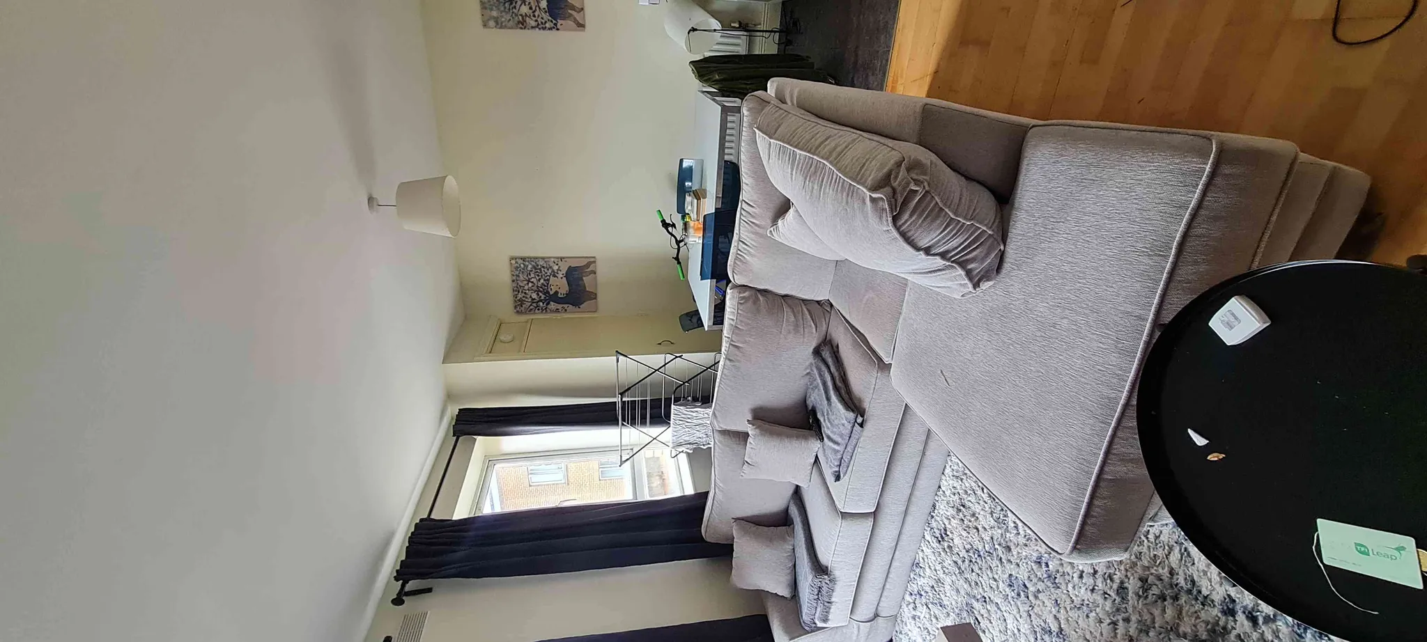 Living room with a large light-gray sectional sofa, round black coffee table, shag rug, curtains and a window; a desk and drying rack visible against the far wall.