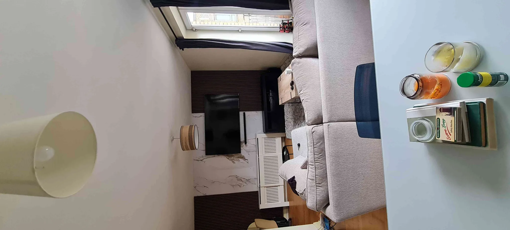 Living room with a light-colored sofa, wall-mounted TV, window with curtains, side table, and a tabletop holding jars and candles.