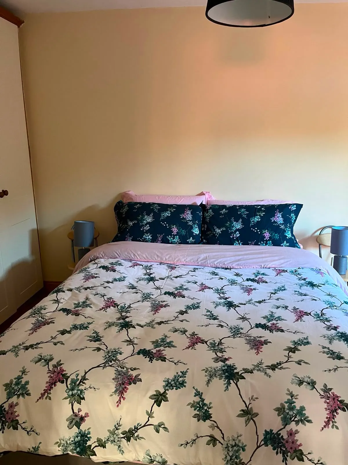 Bedroom with a double bed, floral duvet, two dark floral pillows, bedside tables with lamps and a wardrobe at left.