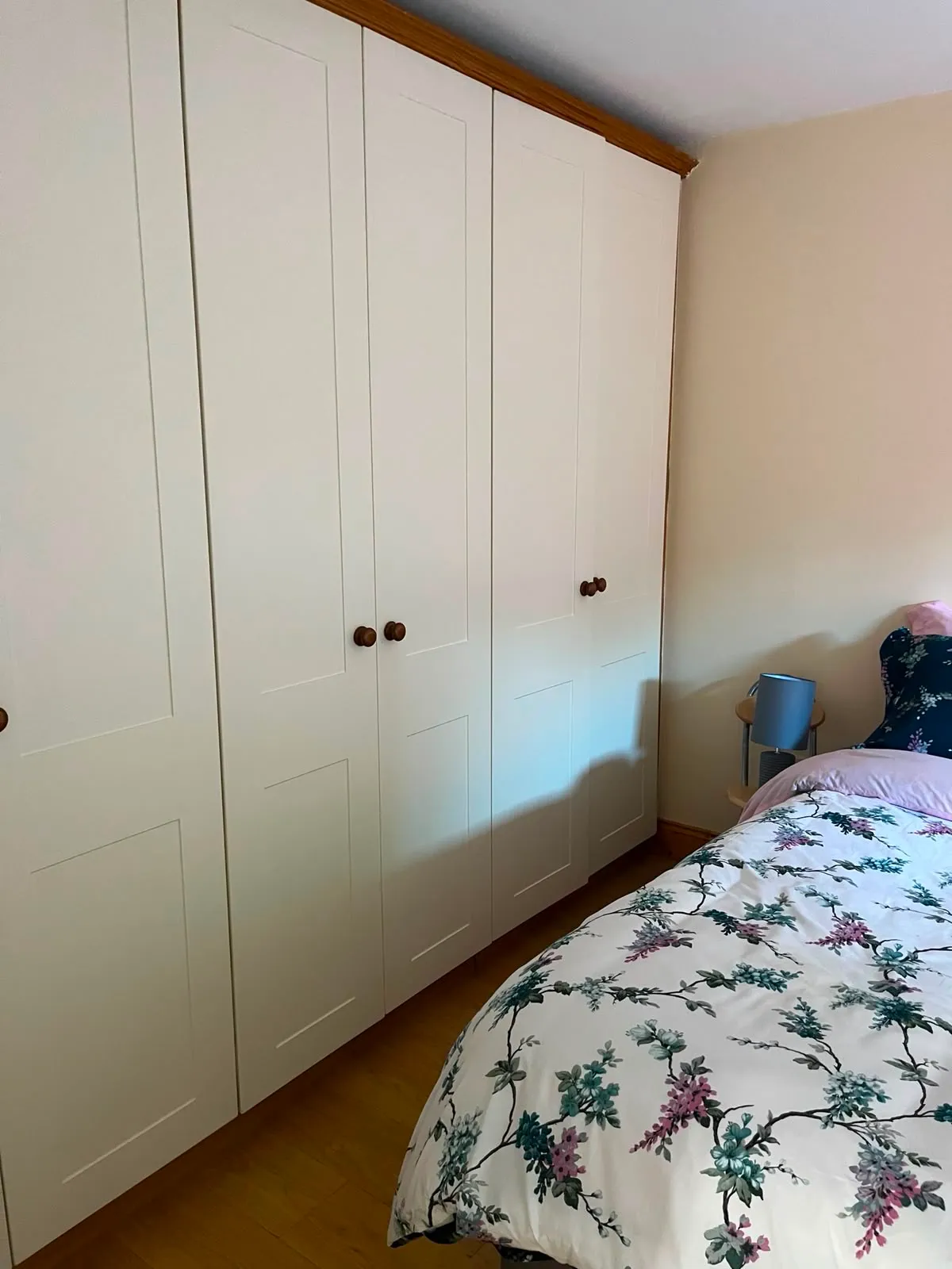 Bedroom corner with a large built-in white wardrobe, a bed with floral duvet and a small bedside table with a lamp.