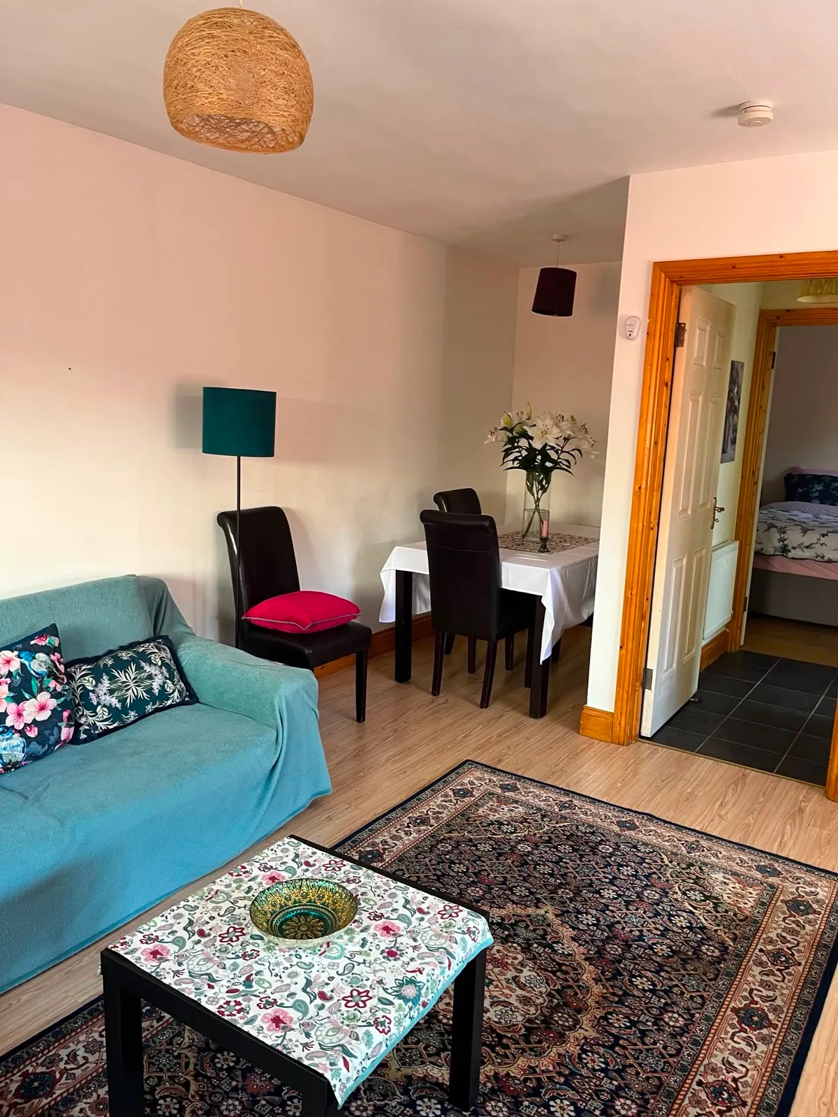 Living room with a sofa, small coffee table, patterned rug, floor lamp and a dining table with chairs; open door shows a bedroom ahead.