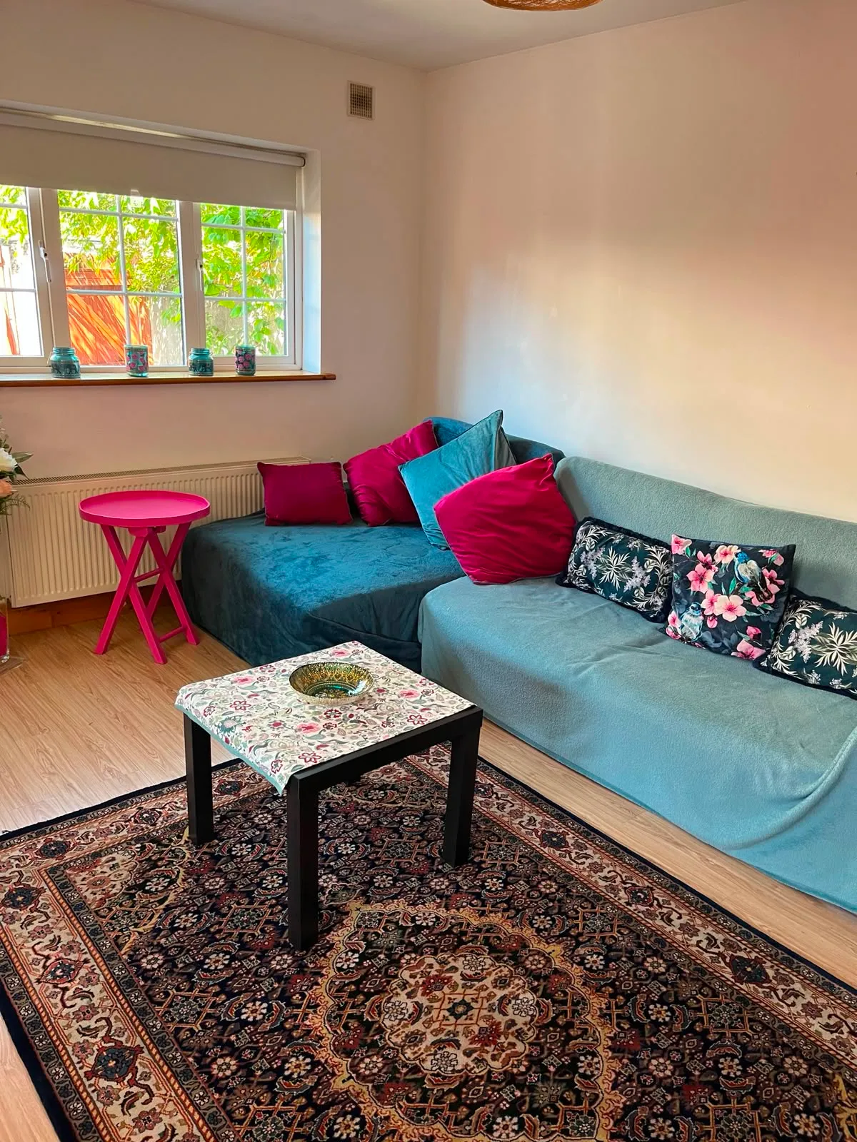 Living room with a blue-covered sectional sofa and colorful throw pillows, small black coffee table on a patterned area rug, window with candles on the sill and a pink side table.