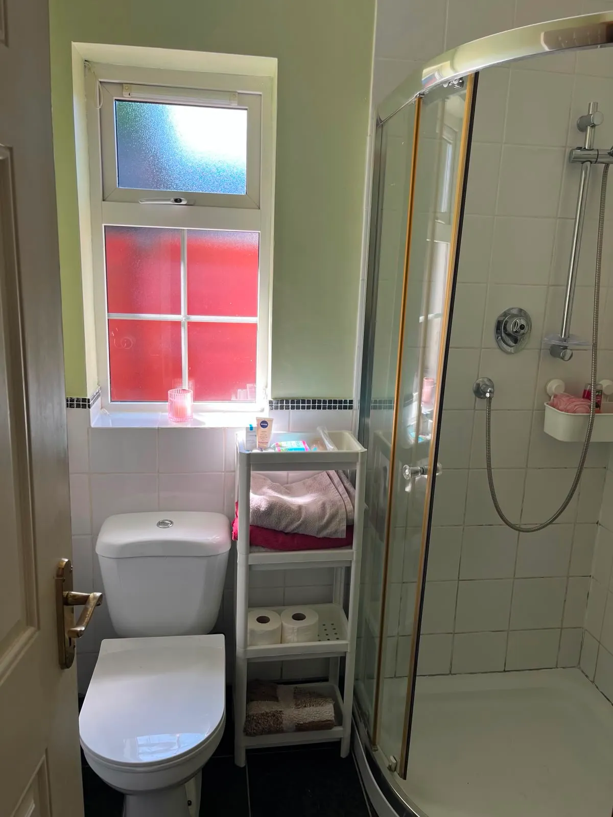 Bathroom with a toilet, a corner glass shower, a frosted window, and a small shelving unit holding towels, toiletries and toilet paper.
