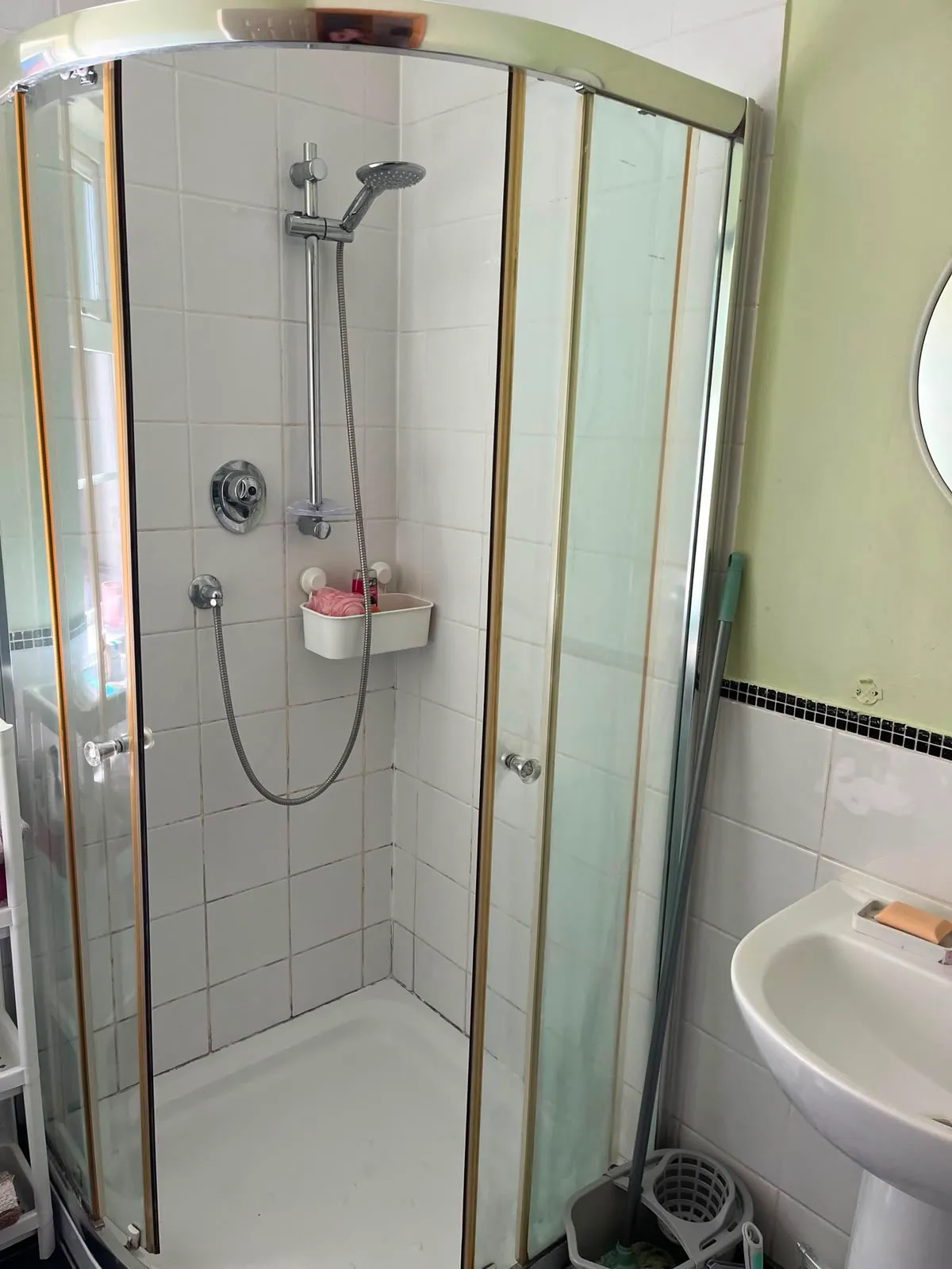 Bathroom with a corner glass shower enclosure, handheld showerhead on a vertical rail, white tiled walls, small wall-mounted soap caddy with a pink cloth, and a pedestal sink partially visible to the right.