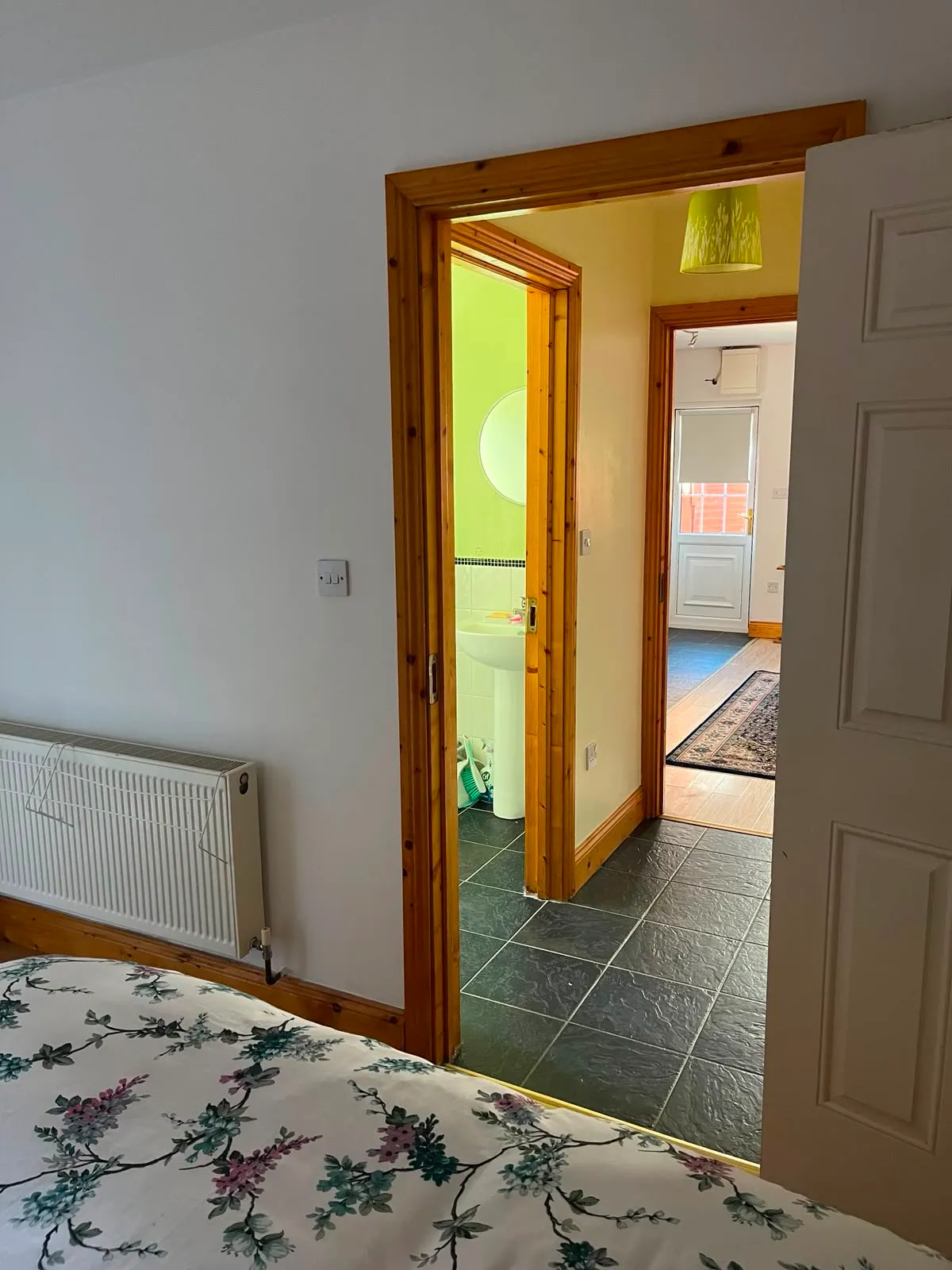Bedroom view showing a floral bed corner, a radiator, and an open doorway leading to a bathroom and hallway