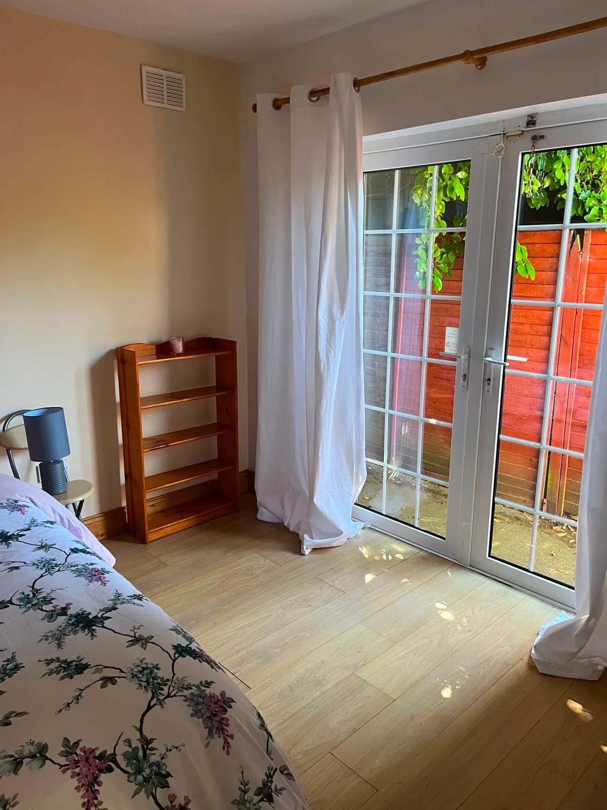 Bedroom corner with a bed edge, small wooden bookshelf, bedside lamp, white curtains and glass double doors to an outside area