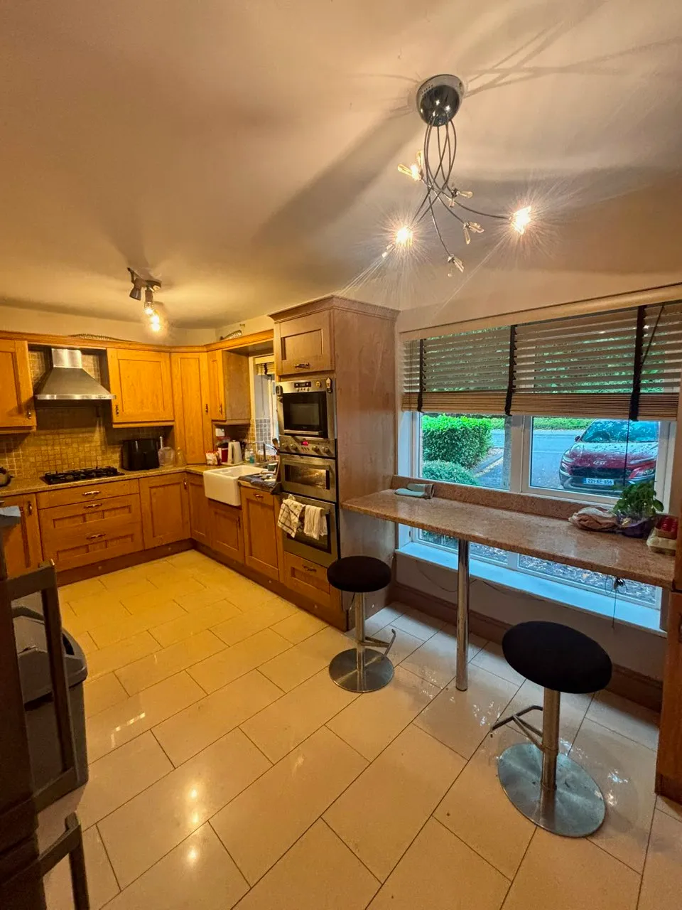 Kitchen with wooden cabinets, built-in oven and hob, sink, breakfast bar with two stools, window with blinds, and overhead lighting