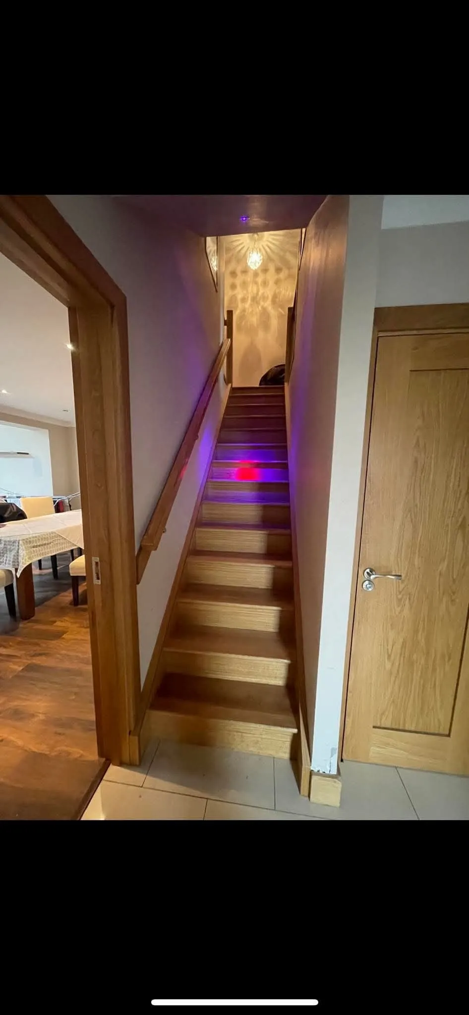 Narrow wooden staircase with handrail leading up to a landing, pendant light casting patterned shadows and colored lights on the steps; doorway and dining area visible to the left.