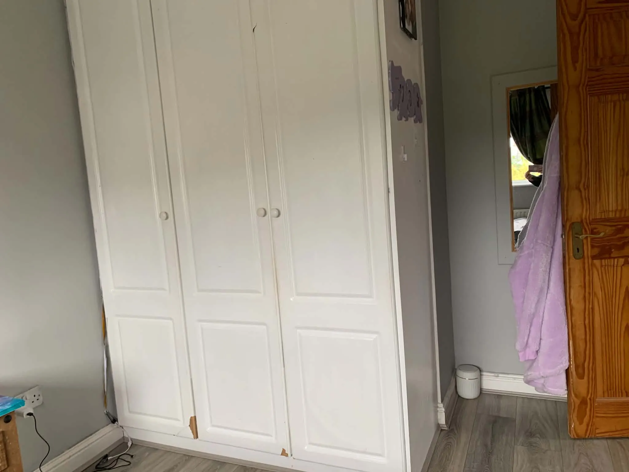 Bedroom corner with a white built-in wardrobe, wooden door with a hanging pink towel, small wall mirror, and laminate floor