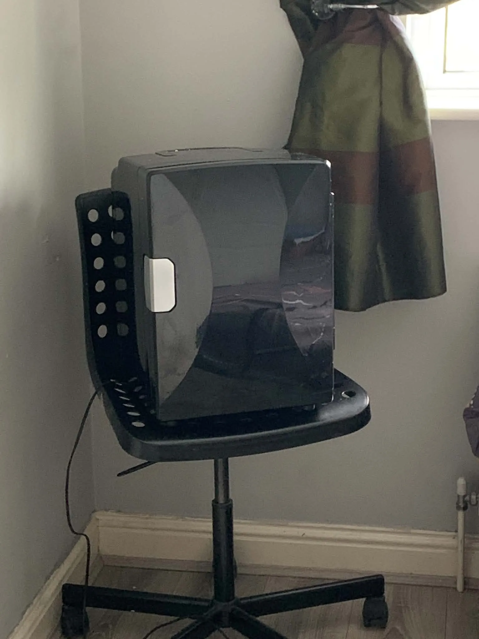 Corner of a room showing a small black appliance on a swivel chair, a green curtain and a window edge, and a visible power cord along the baseboard.