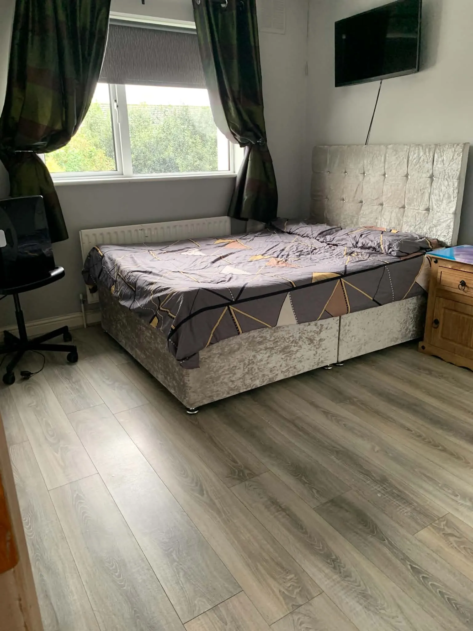 Bedroom with a double bed, padded headboard, window with curtains, wall-mounted TV and a wooden bedside cabinet.